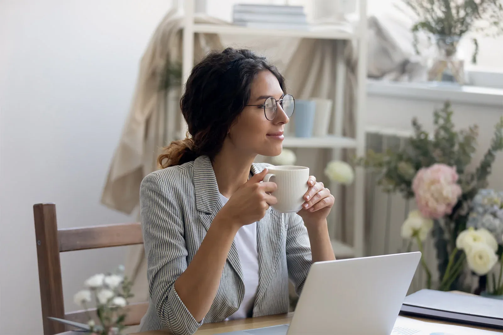 A woman, wearing glasses, sits at a desk in a home office, thoughtfully holding a mug. A laptop sits in front of her.