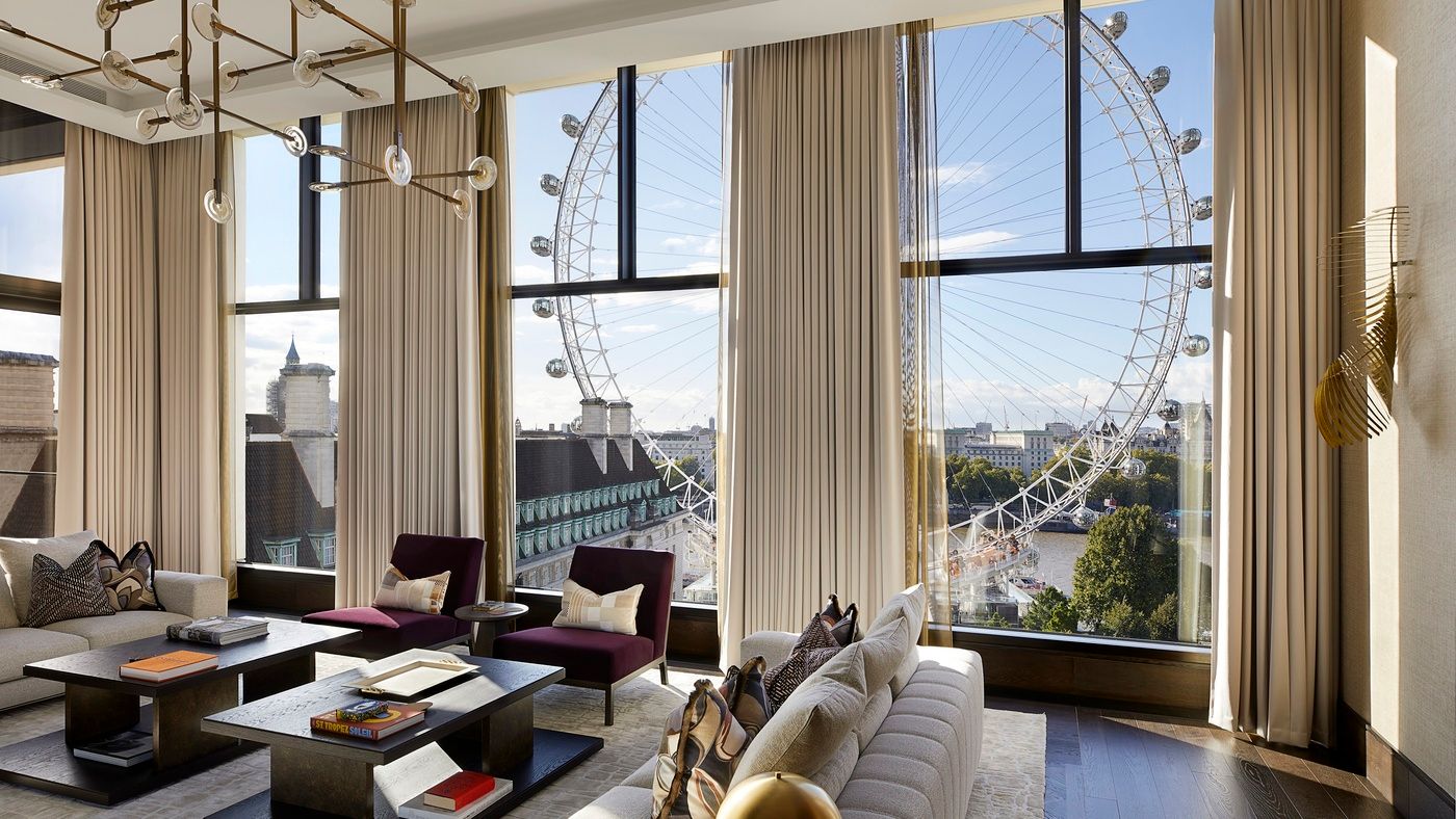 A living room overlooks the London Eye. Beige curtains frame floor-to-ceiling windows showcasing the Ferris wheel and city skyline. Two armchairs and a sofa are arranged around coffee tables.
