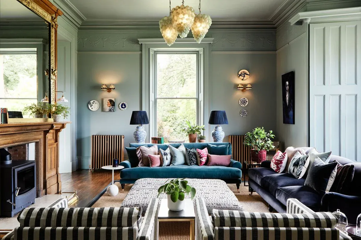 A teal velvet sofa sits in a spacious, sage-green room. It is adorned with numerous pillows and flanked by dark velvet sofas. A patterned ottoman rests in front. The room features large windows, ornate molding, and various decorative objects.