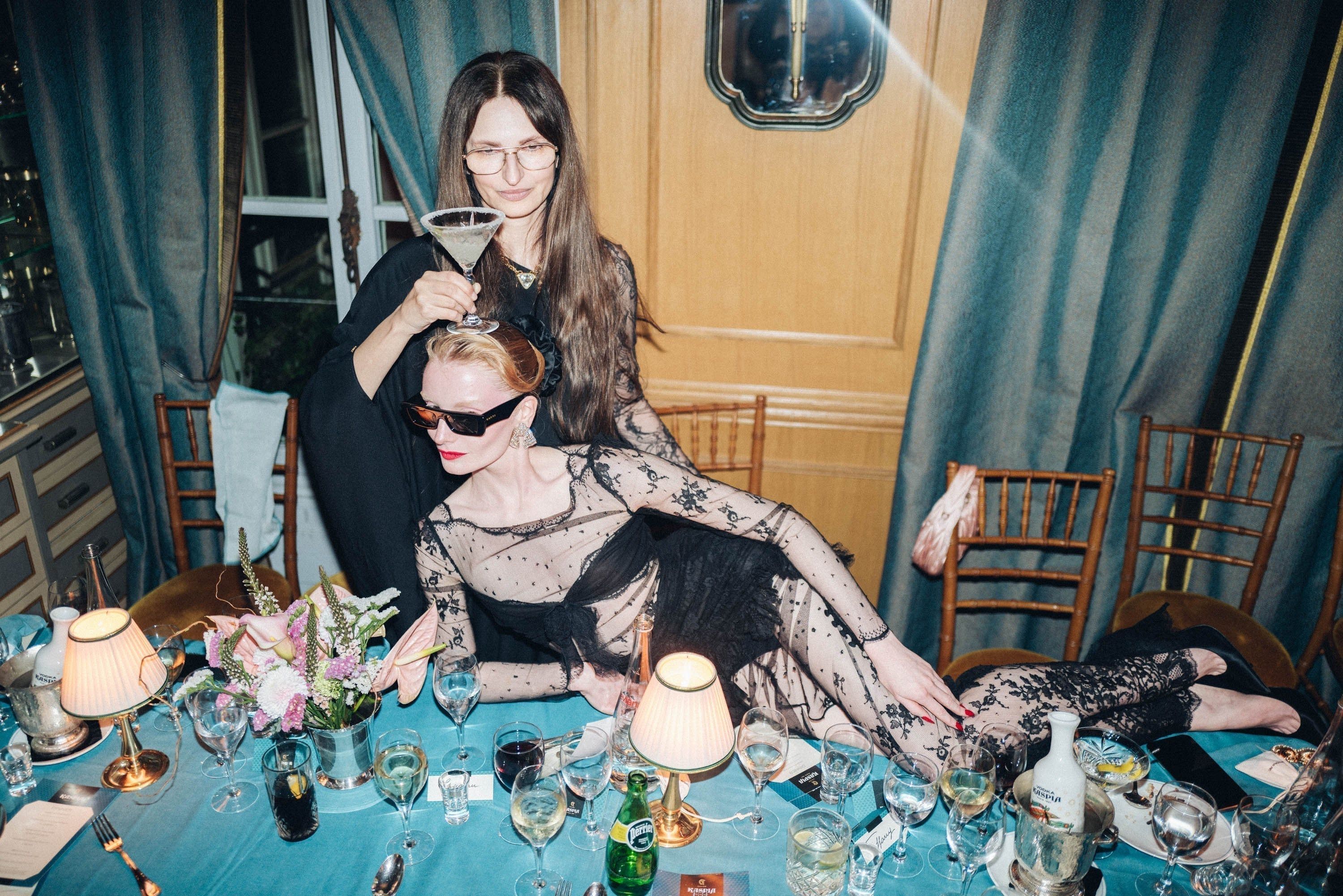 Two women, one placing a cocktail on the other's head, recline playfully at a formal dinner table. The table is set with glasses, bottles, and a floral arrangement.