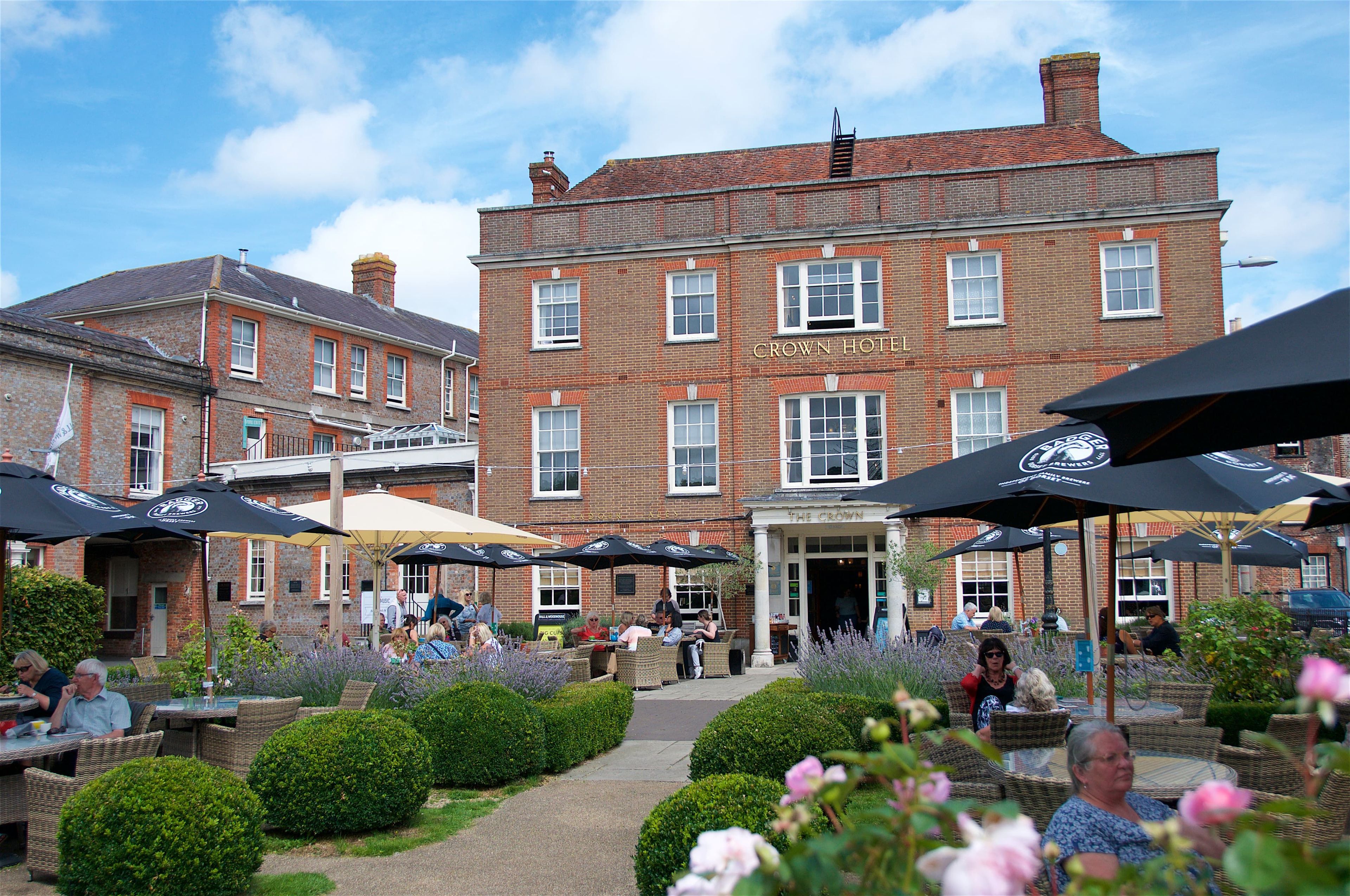 A brick Crown Hotel sits beyond a patio with patrons at tables under umbrellas. Lavender and hedges border the walkway. CROWN HOTEL is visible above the entrance.