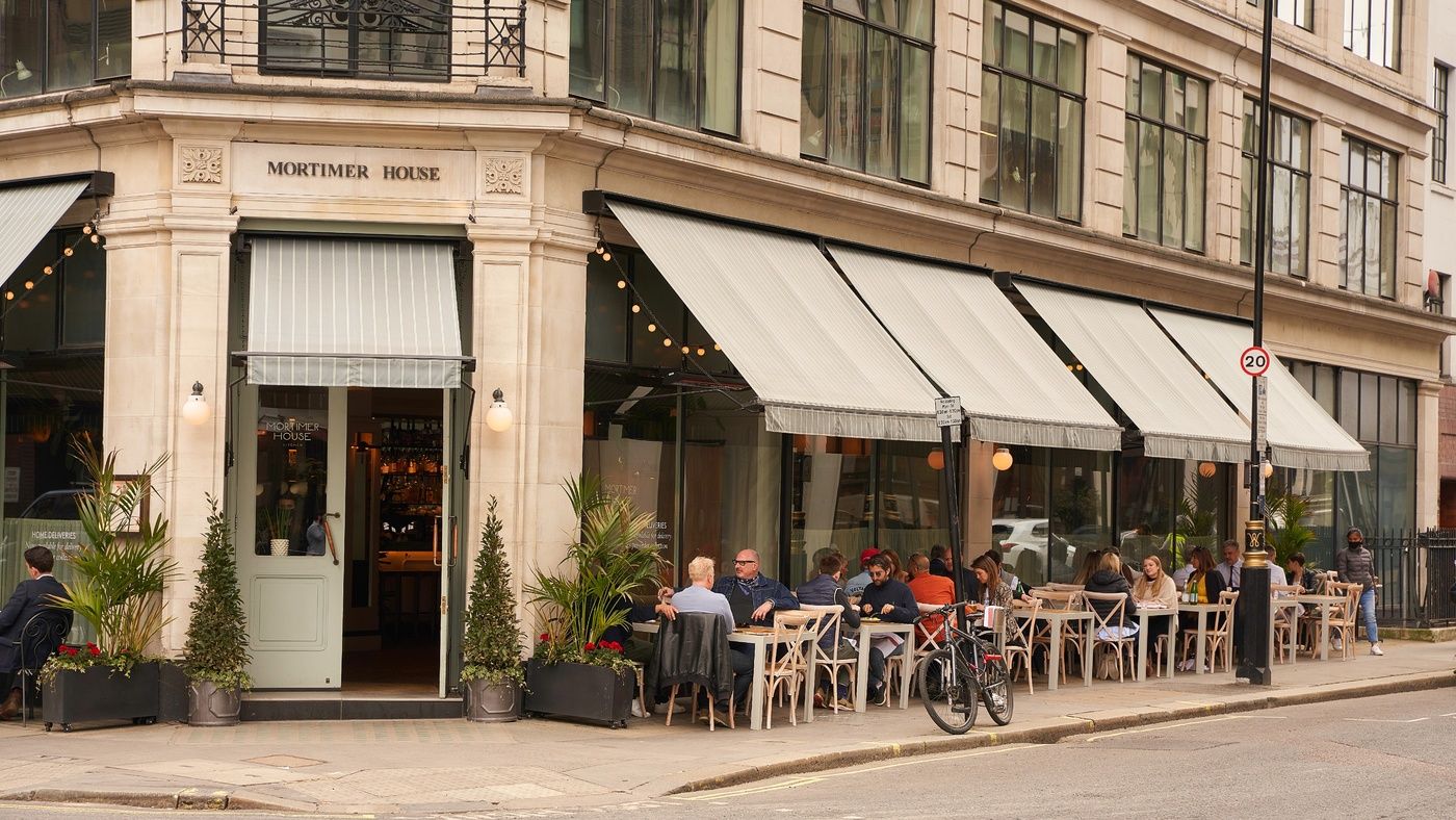 A restaurant, Mortimer House, with patrons dining outside at tables under striped awnings on a city street.