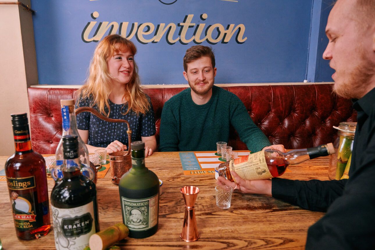 Two people sit on a red leather banquette, a bartender pours liquor. The wall behind them features the word "invention".