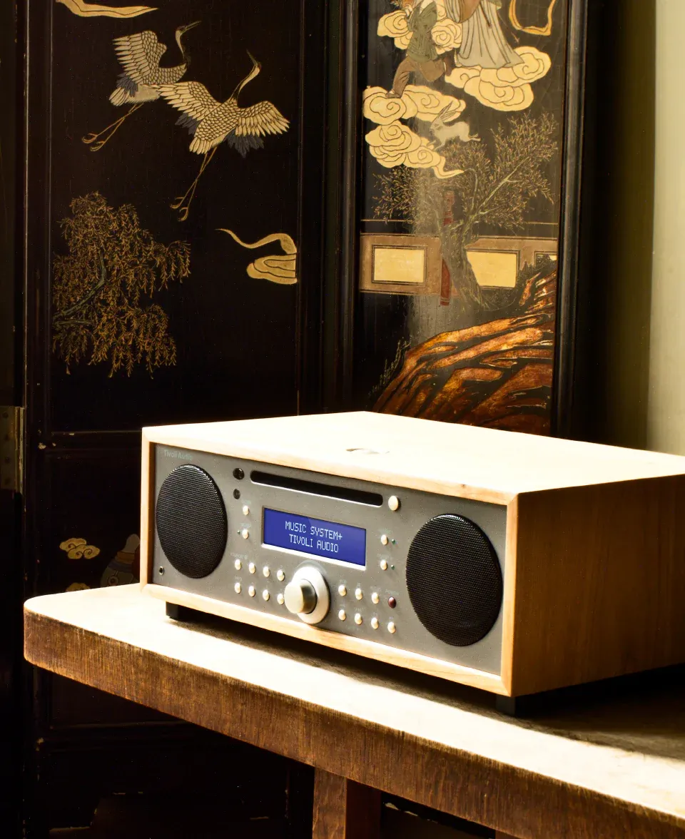 "A Tivoli Audio music system sits on a wooden table, displaying ""MUSIC SYSTEM+ TIVOLI AUDIO"" on its screen. It rests in front of an ornate, dark-wood screen.
