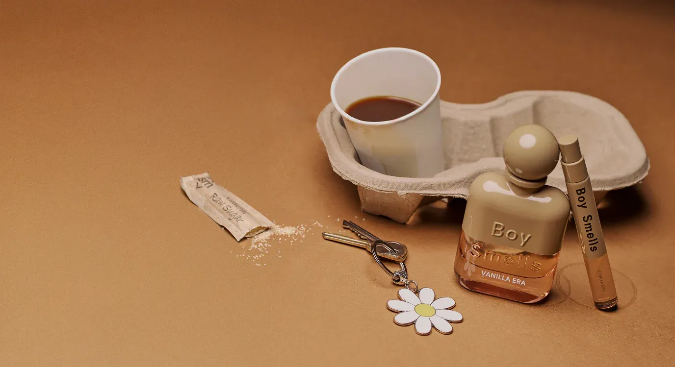 A Boy Smells Vanilla Era perfume and its travel-size counterpart rest on a cardboard coffee cup holder. A coffee cup, raw sugar packet, and keys with a daisy keychain are nearby on a brown surface.