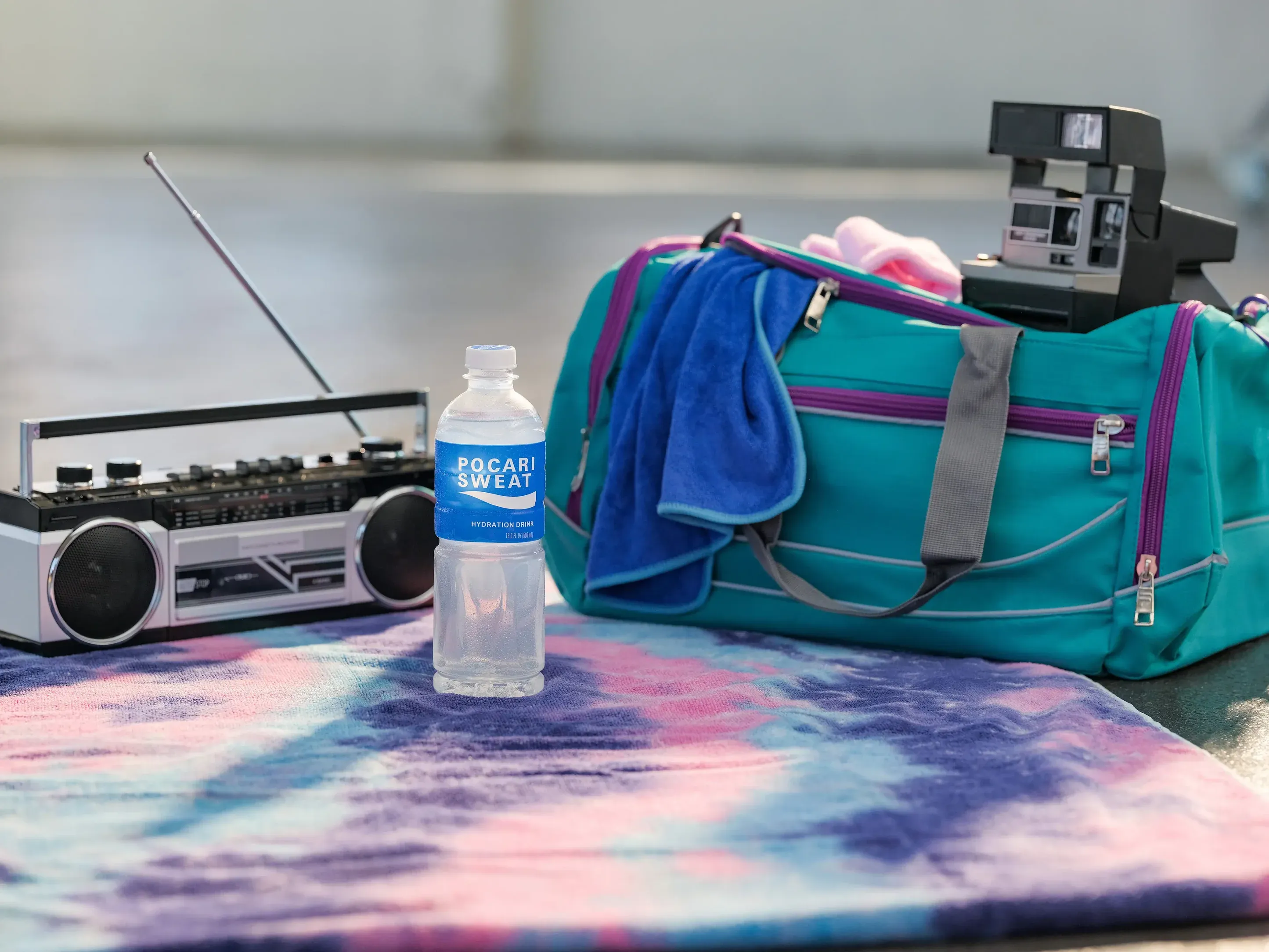 A Pocari Sweat hydration drink bottle rests on a tie-dye towel near a boombox, teal gym bag, and an instant camera.
