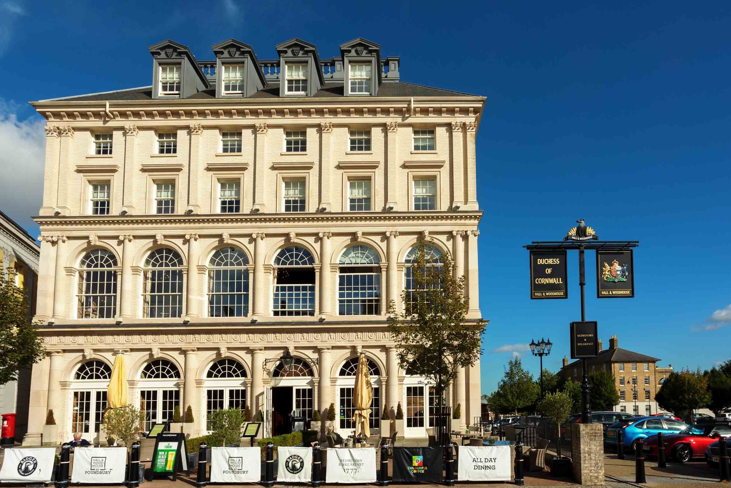 A large, tan building with many arched windows sits in a town square. The building is a pub called ""Duchess of Cornwall,"" with outdoor seating. Cars are parked nearby.