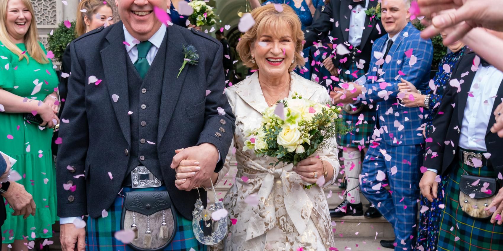 A bride and groom, holding hands, exit a building showered in confetti. The bride carries a bouquet. The groom wears a kilt.