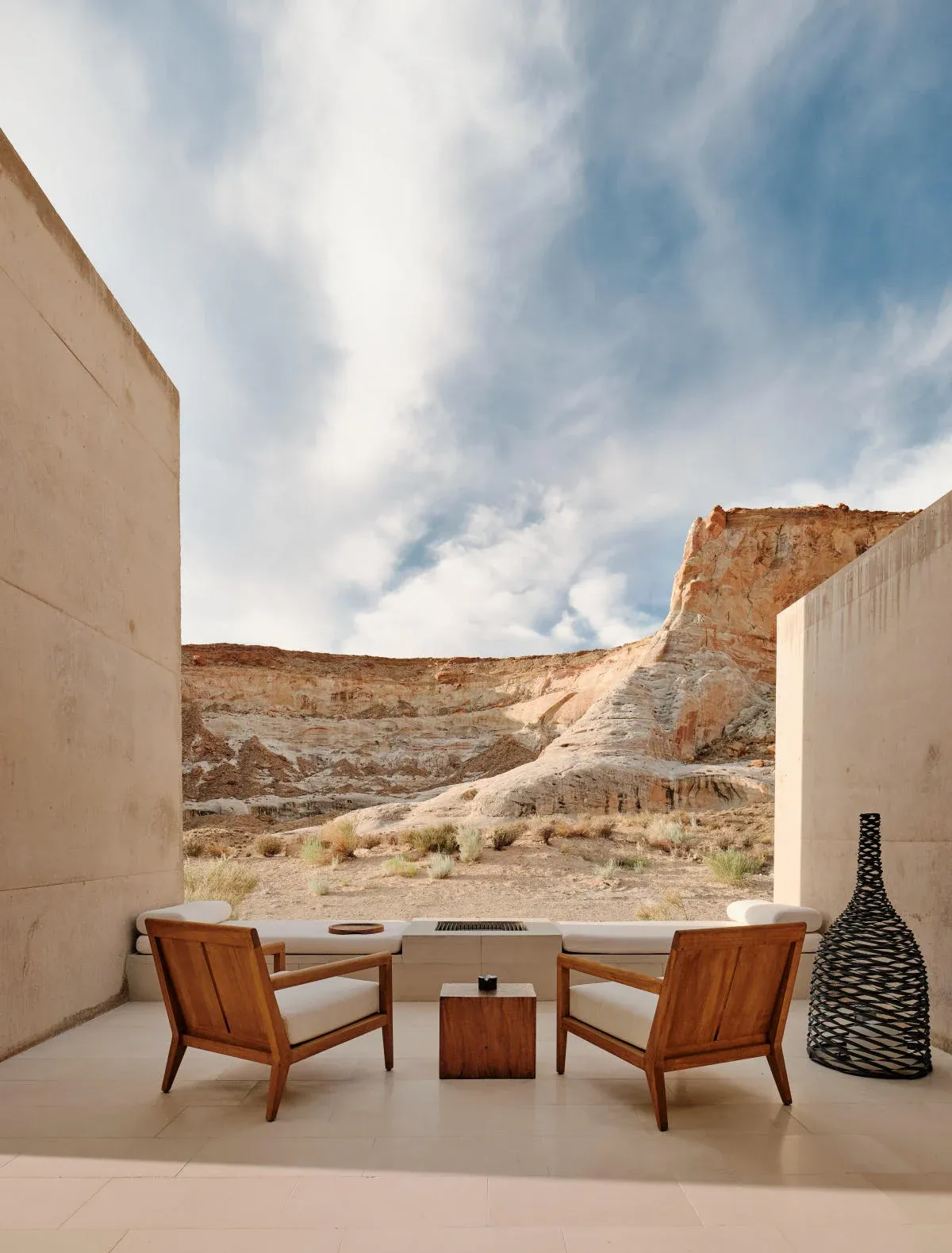 Two wooden chairs sit on a patio, facing a desert landscape with layered rock formations. A small wooden table is between them. A woven vase stands near a concrete wall.