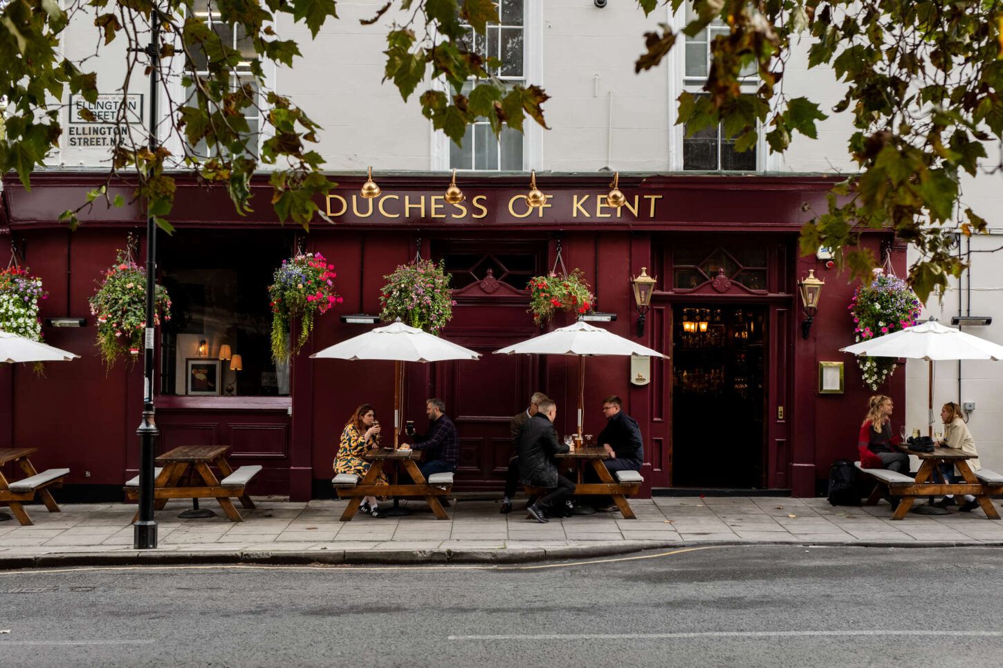 A maroon pub, the "Duchess of Kent," displays hanging flower baskets. Patrons sit at outdoor picnic tables under white umbrellas. The pub is located on a city street, partially obscured by overhanging tree branches.